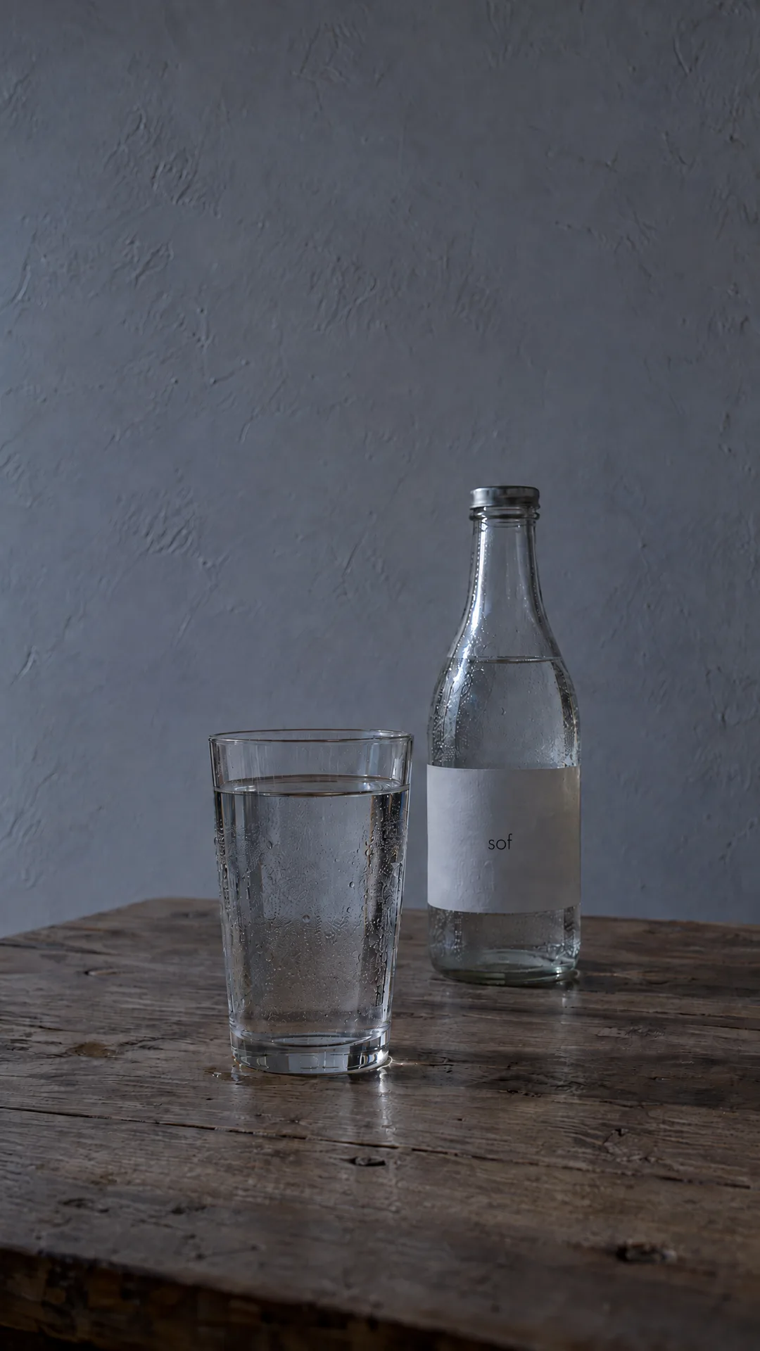 A glass of water and a bottle labelled sof on a rustic oak table at dusk